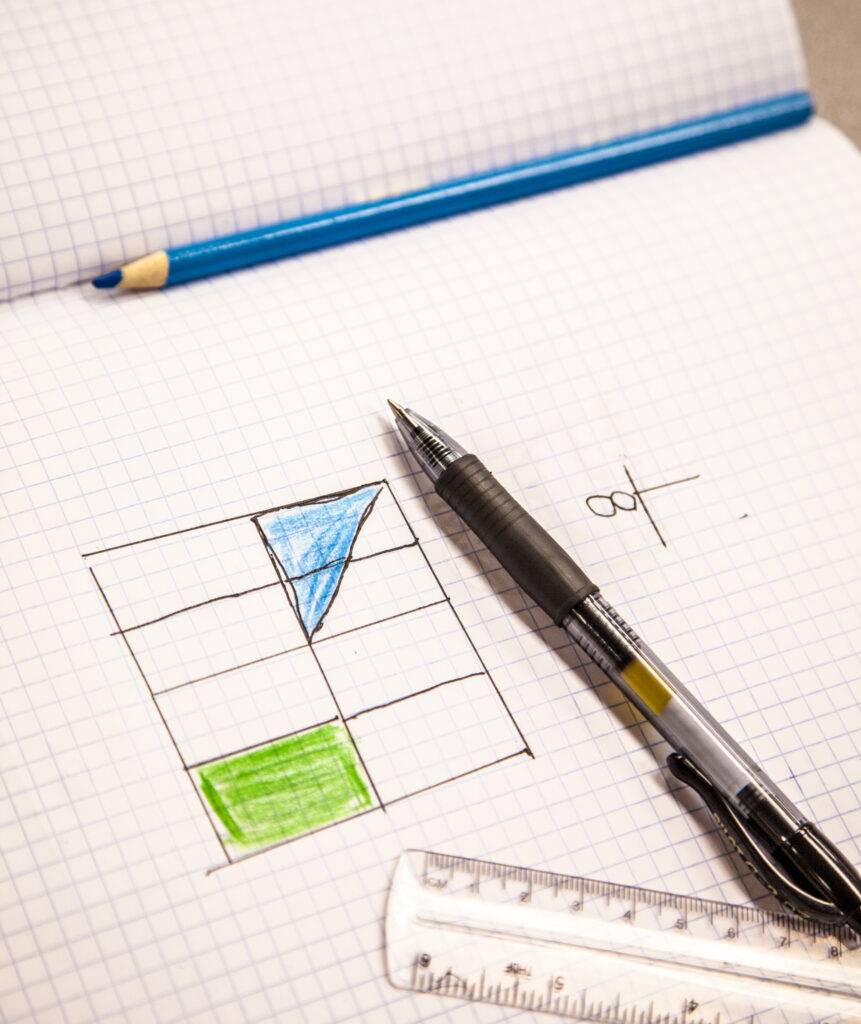A top-down view of a graph paper notebook featuring hand-drawn geometric shapes colored in blue and green. A black pen, a blue colored pencil, and a clear plastic ruler are resting on the pages.